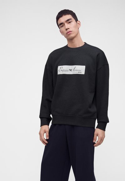 Άνδρας που φοράει μαύρο φούτερ Emporio Armani και σκούρο παντελόνι, στέκεται μπροστά σε απλό ανοιχτογκρι φόντο.