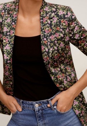 Osoba ubrana w czarną górę, niebieskie dżinsy i marynarkę w kwiatowy wzór z różowymi i zielonymi kwiatami, ręce spoczywające w przednich kieszeniach.