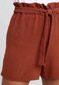 Szorty w kolorze rdzawego brązu wykonane z teksturowanej tkaniny, z marszczoną talią i wiązanym paskiem dla regulowanego dopasowania.