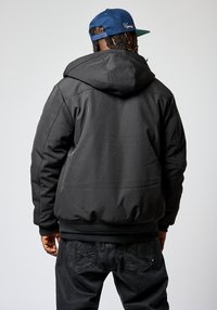Chaqueta acolchada negra con capucha, que presenta detalles de costura visibles y puños elásticos. Combinada con unos jeans oscuros acentuados por un patrón texturizado.