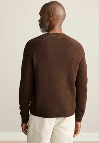 Brauner Strickpullover mit rundem Hals, gerippten Bündchen und Saum; hat eine strukturierte Oberfläche und Raglanärmel. Lässige Passform, getragen mit hellen Hosen.
