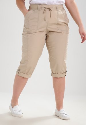 Personne portant un pantalon beige mi-longueur avec taille à cordon et revers roulés, associé à des chaussures blanches sans lacets, se tenant devant un fond uni.