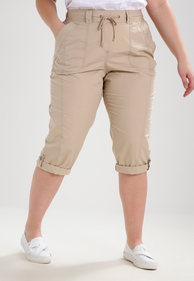 Personne portant un pantalon beige mi-longueur avec taille à cordon et revers roulés, associé à des chaussures blanches sans lacets, se tenant devant un fond uni.