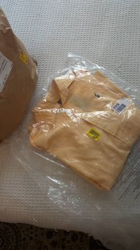 Camisa às riscas amarelas em embalagem plástica, com gola e botões. Inclui uma etiqueta de preço visível. Embalagem castanha ao fundo.