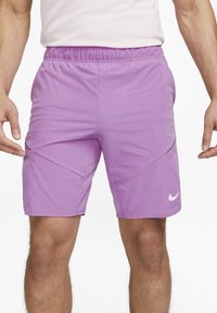 Man i ljuslila Nike träningsshorts med elastiskt midjeband och vit Nike-logotyp på vänster lår, stående mot vit bakgrund.