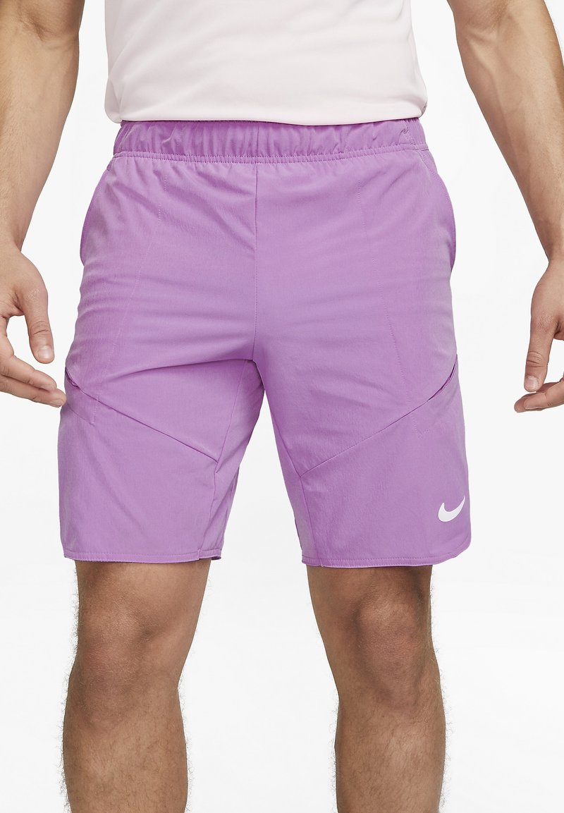 Man i ljuslila Nike träningsshorts med elastiskt midjeband och vit Nike-logotyp på vänster lår, stående mot vit bakgrund.