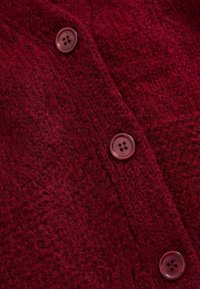 Texturovaná burgundská pletenina s mírně chlupatým povrchem. Čtyři odpovídající knoflíky s kulatým, hladkým designem.