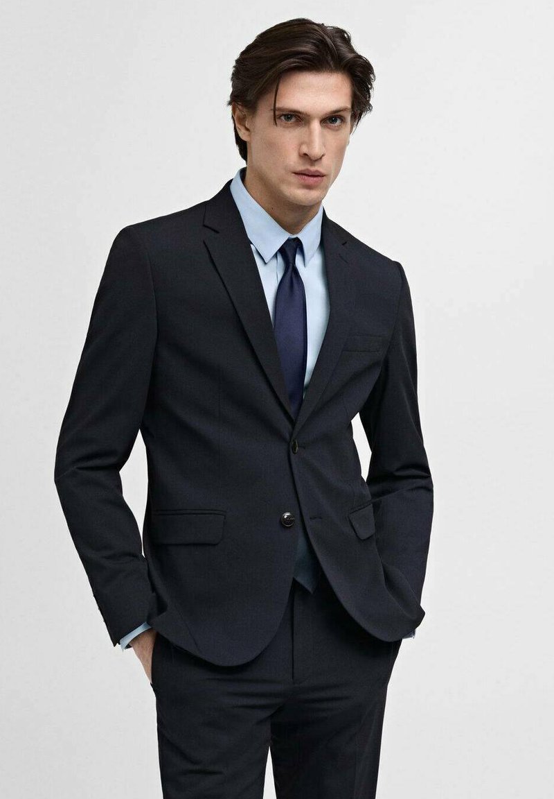 Costum bleumarin cu rever înclinat și două buzunare frontale, asortat cu o cămașă de dress de un albastru deschis și o cravată închisă. Material neted, croială ajustată.
