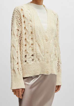 Femme portant un cardigan épais en maille crème avec des motifs torsadés et ajourés, par-dessus un haut blanc et une jupe en satin taupe clair.