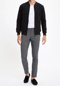 Czarna teksturowana kurtka bomberka na białej koszulce, w połączeniu z dopasowanymi szarymi spodniami w kratę i czarnymi loafersami.