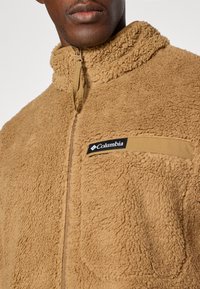 Beige fleecejack met een hoge kraag, halve rits aan de voorkant en een borstzak met een zwart Columbia-logo. Zacht, gestructureerd materiaal.