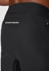 Crne atletske gaće s glatkom teksturom, visokim pojasom, bočnom patentni zatvaračem i bijelim logotipom "Sweet Protection" na stražnjoj strani.