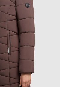 Jachetă pufoasă maro cu textură matlasată, având o inscripție neagră pe mânecă și un buzunar lateral cu fermoar.