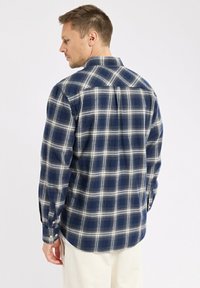 Homme portant une chemise à carreaux bleue et blanche à manches longues, tourné sur le côté, avec un pantalon de couleur claire devant un fond uni.