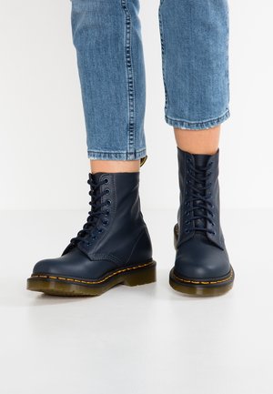 Mørkeblå læderstøvler med gule syninger og snørebånd, brugt med korte blå jeans på en hvid gulv.