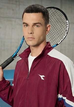 Ung mann i en burgunder og hvit sportsjakke holder en tennisracket over skulderen, står inne ved en vegg.