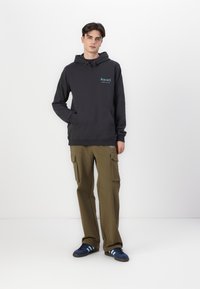 Dunkelgrauer Kapuzenpullover mit Fronttasche und türkisfarbenem Logo, kombiniert mit olivgrünen Cargo-Hosen mit mehreren Taschen und Reißverschlüssen.
