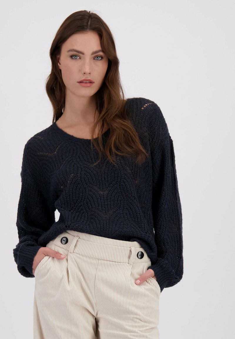 Vrouw met lang bruin haar, gekleed in een donkerblauwe gebreide trui en crèmekleurige corduroy broek met hoge taille, met de handen in de zakken.