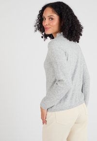 Maglione grigio chiaro con una texture morbida, maniche lunghe e un collo alto, abbinato a pantaloni color crema. Design liscio e minimalista.