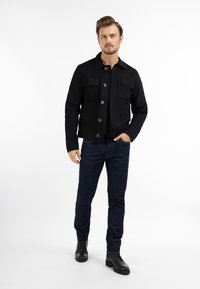 Sort bomuldsjakke med frontlommer og knaplukning, parret med mørkeblå jeans og sorte læderstøvler mod en hvid baggrund.