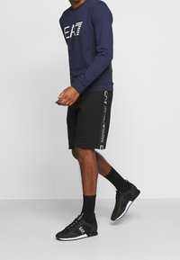 Navy sweatshirt med vit "EA7" logga, svarta shorts med loggaakcentueringar och svarta träningsskor med vit sula. Avslappnad sportig outfit.