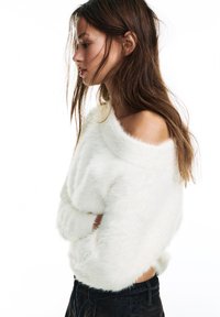 Młoda kobieta z długimi brązowymi włosami nosi biały puszysty sweter odsłaniający ramiona oraz czarne spodnie, pozując z profilu na białym tle.
