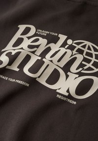 Tessuto scuro con grandi scritte audaci in grigio chiaro, con la scritta "Berlin STUDIO", testo aggiuntivo sopra e sotto, e una grafica di un globo.
