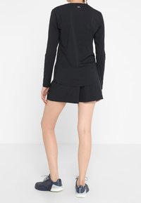 Female wearing black long-sleeve athletic top, black shorts, and navy running shoes, standing against a white plain background, back facing viewer.