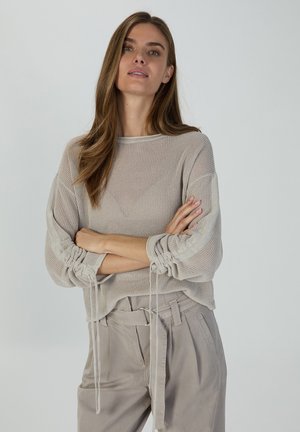 Light beige knitted sweater with a loose fit, featuring cinched sleeves and pull ties. Worn with light gray trousers, showcasing a relaxed style.