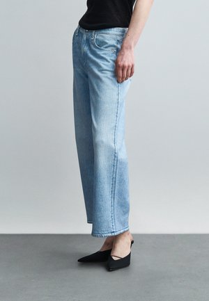 Άτομο που φορά ανοιχτό μπλε τζιν με ίσια γραμμή, μαύρες γόβες με μυτερή μύτη και μαύρο τοπ, στέκεται πάνω σε γκρι πάτωμα μπροστά σε απλό φόντο.