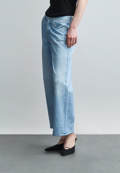Person wearing light blue straight-leg jeans, black pointed-toe heels, and a black top, standing on a gray floor against a plain backdrop.