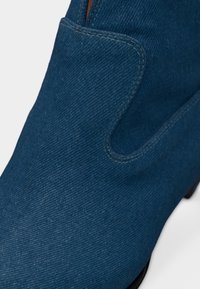 Κοντινό πλάνο ενός μπλε τζιν μποτακιού που δείχνει λεπτομερή ραφές και σχεδιασμό με ανοίγματα στο πάνω μέρος.