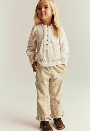 Hvid, struktureret langærmet top med knapper og broderet blomstermotiv, kombineret med beige bukser med små blomsterprint og flæsekantede manchetter.