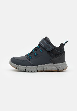 Baskets de sport avec une tige synthétique noire, des accents bleus, une sangle à boucle et une semelle en caoutchouc gris texturé avec des détails mouchetés.