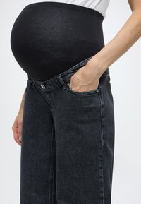Τζιν εγκυμοσύνης με μαύρη ελαστική μέση, ύφασμα σκούρου τζιν, φαρδιά γραμμή και μπροστινές τσέπες. Μια χέρι ακουμπά στη μέση.