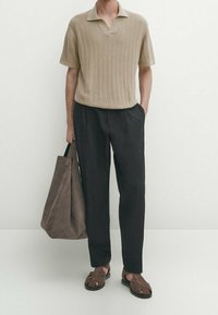 Homme portant un polo en maille beige à manches courtes, un pantalon noir, des sandales marron, tenant un grand sac fourre-tout en daim marron avec une main dans la poche.