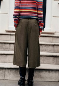 Isik, kes kannab oliivirohelisi laia lõikega lühikesi pükse, musti saapaid ja värvilist triibulist pika varrukaga särki, seisab kivist treppidel.