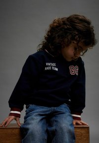 Tumedat sinise värvusega dressipluus valgete "VINTAGE RACE TEAM" kirjutiste ja numbri "86"-ga, mis on ühendatud helesiniste teksade ja tumepunaste aksentidega varrukatega.