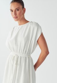 Valkoinen pellavamekko, jossa on venepääntie, lyhyet hihansuut, rypytetty elastinen vyötärö ja kiristysnauha. Suora tekstuuri ja rento malli.