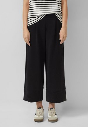 Osoba ubrana w czarne spodnie z szerokimi nogawkami za kostkę, w paskowaną koszulę wpuszczoną w spodnie oraz białe i beżowe trampki, stojąca na tle jasnego, jednolitego tła.