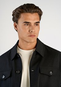 Svart jacka med krage, knappdetaljer och två bröstfickor, bärs över en ljusfärgad t-shirt med rund halsringning. Slät textur, skräddarsydd passform.