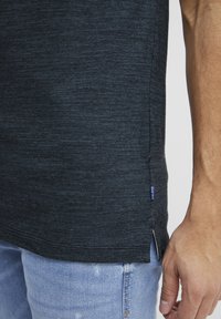 Chemise courte à manches en gris foncé, fabriquée dans un tissu texturé avec des fentes latérales. Présente une petite étiquette logo bleue près de l'ourlet. Portée avec un jean en denim clair.