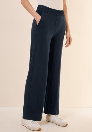 Pantalons larges bleu marine avec une taille élastique, deux poches latérales et une texture lisse, associés à des baskets de couleur claire.