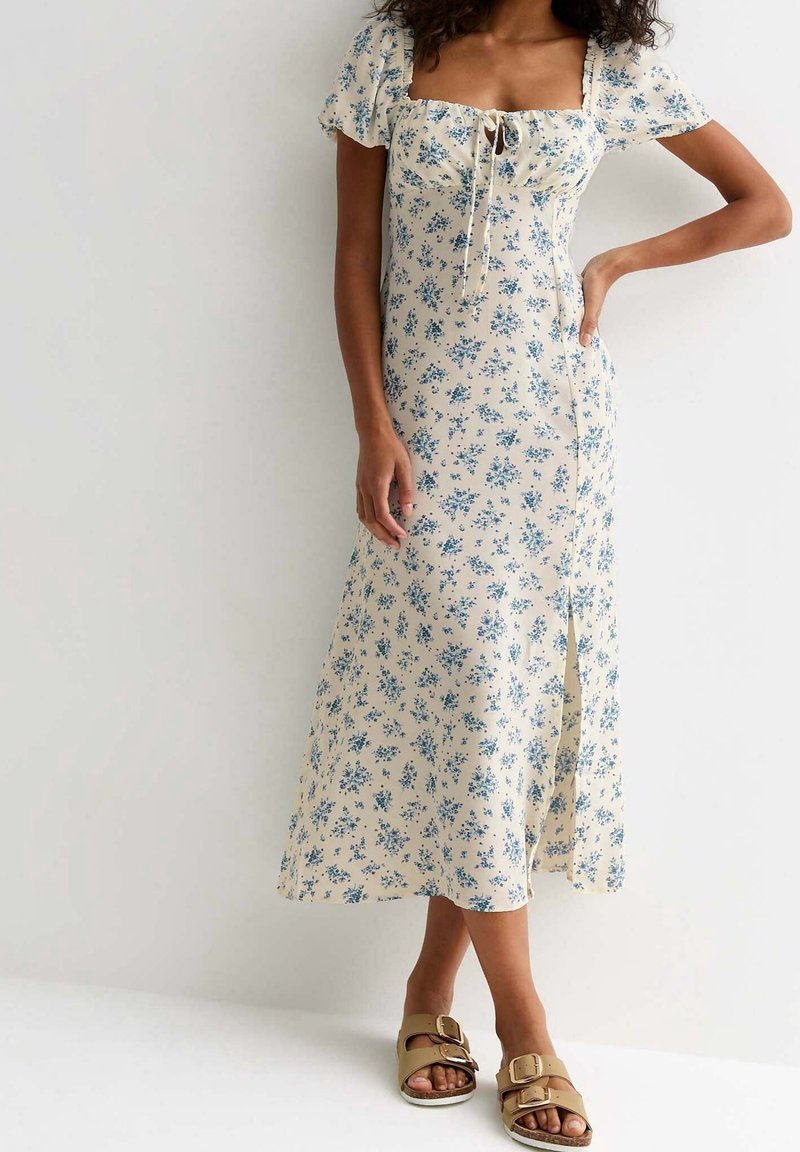 Frau trägt ein cremefarbenes Midikleid mit blauem Blumenmuster, Puffärmeln und Schlitz vorne, kombiniert mit beigefarbenen Sandalen mit Schnallen.