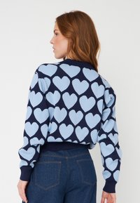 Sweater azul-marinho com padrões de corações azul-claro, punhos e faixa na cintura canelados. Tecido macio com um corte relaxado e design texturizado.
