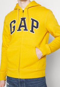 Sudadera con capucha amarilla con cremallera, confeccionada en tela suave, que presenta un gran logo "GAP" en azul marino en el frente, capucha ajustable y bolsillos delanteros.