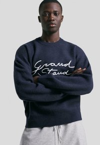 Mlad črn moški stoji z crossed arms, oblečen v temnobluzo s belim kurcivnim napisom "Grand Stand" in svetlo sivo hlače, na enobarvni ozadju.