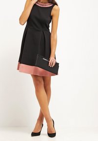 Svart klänning med en figurnära livstycke, en utsvängd kjol och en rosa fåll. Åtföljd av en svart clutch och svarta högklackade skor. Smyckad halsringning.