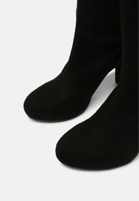 Paire de bottes mi-mollet en daim noir avec bouts arrondis et talons blocs sur un fond uni clair.