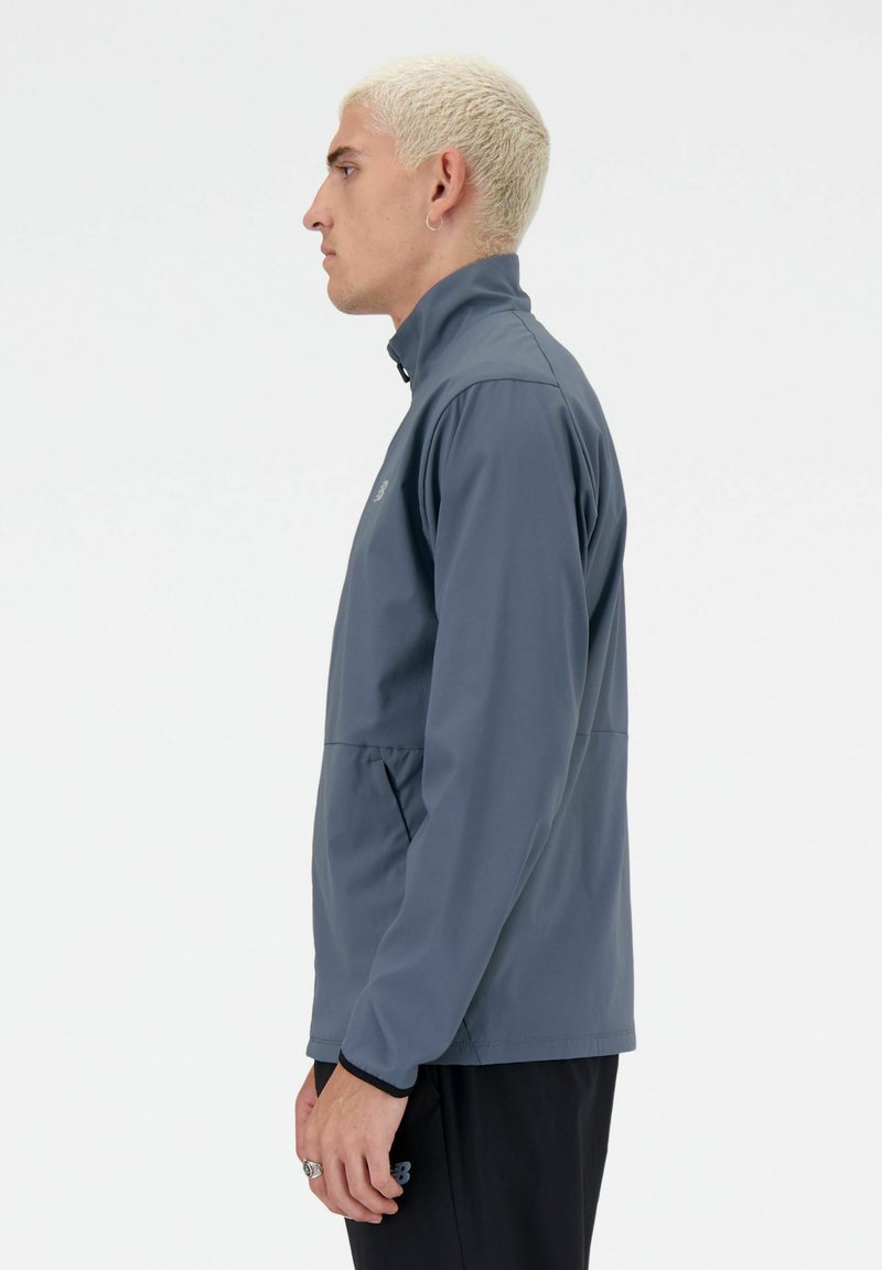 Joven con el cabello corto y decolorado lleva una chaqueta azul marino con cremallera y pantalones negros, de pie de perfil contra un fondo blanco liso.